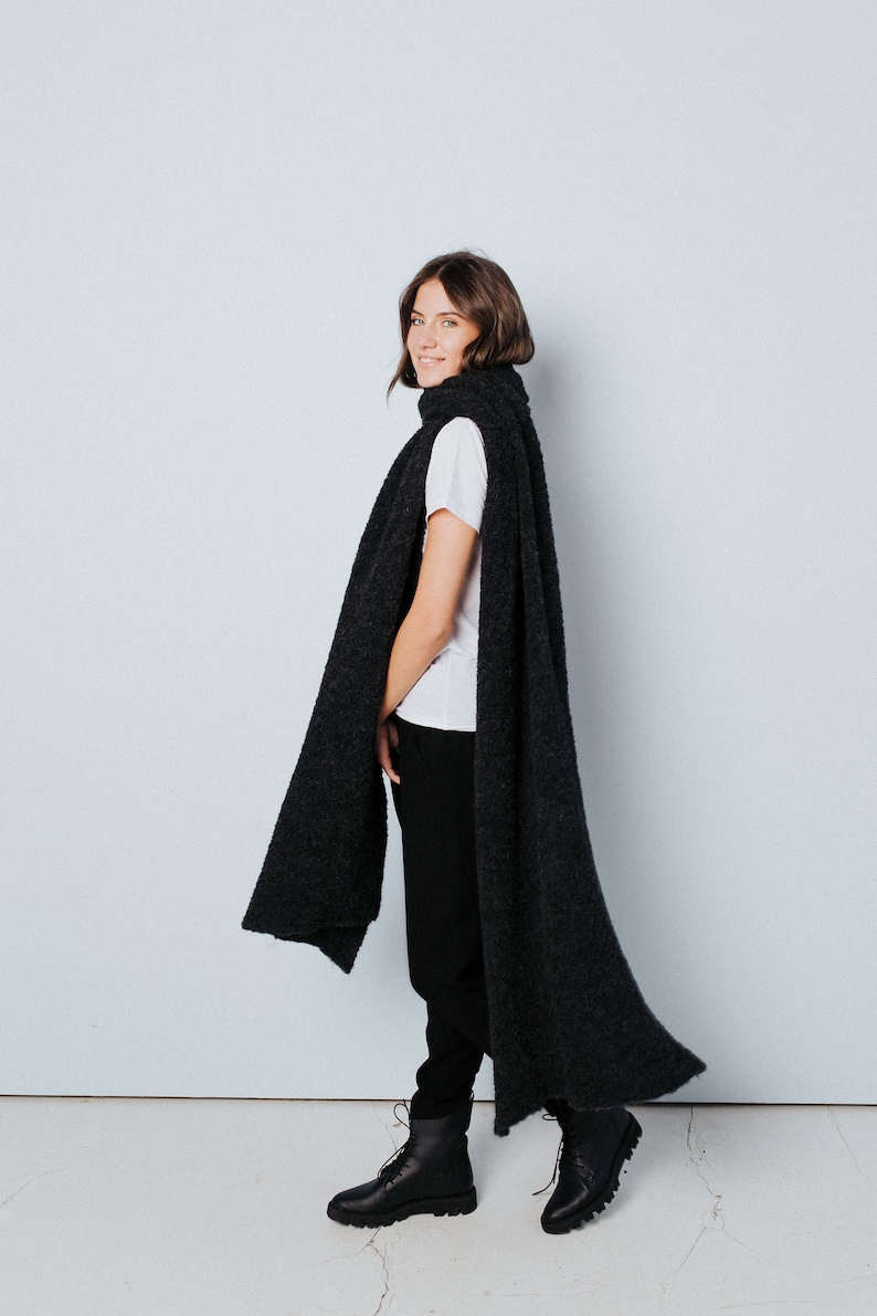 May include: A woman wearing a black fuzzy scarf, a white t-shirt, black pants, and black boots. She is standing in front of a light blue wall.