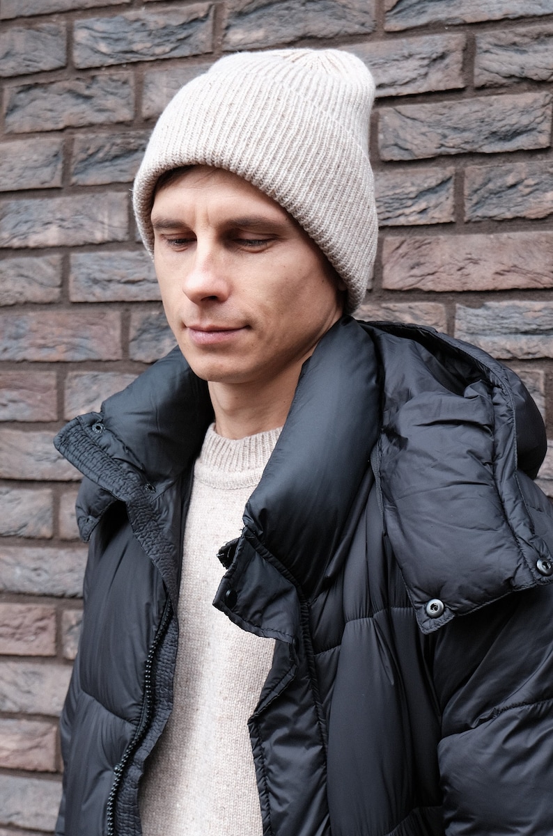 K&ouml;nnte beinhalten: Ein Mann tr&auml;gt eine beige Strickm&uuml;tze und eine schwarze Daunenjacke mit Kapuze. Die Jacke hat einen Rei&szlig;verschluss und Kn&ouml;pfe.