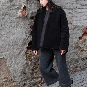 K&ouml;nnte beinhalten: Eine Person tr&auml;gt eine schwarze Fleecejacke, eine schwarze M&uuml;tze und eine graue, weite Hose. Sie steht neben einer Backsteinmauer mit abgeplatztem Putz.
