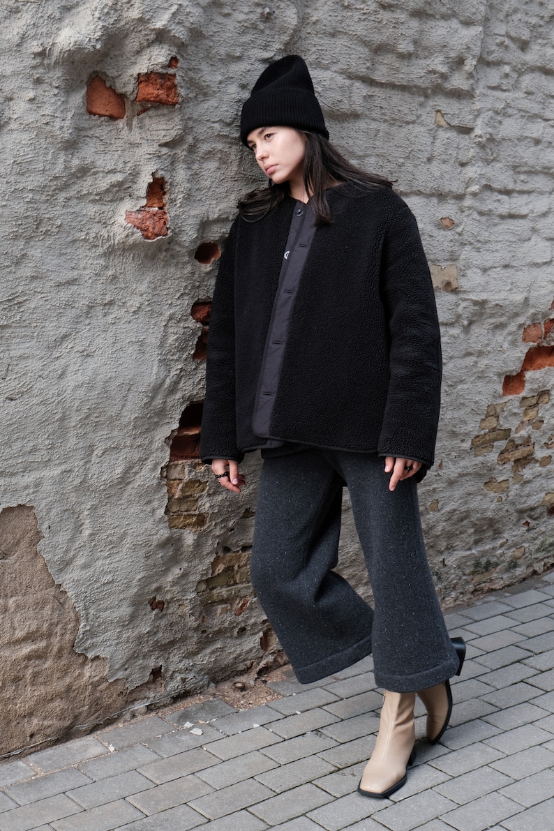 K&ouml;nnte beinhalten: Eine Person tr&auml;gt eine schwarze Fleecejacke, eine schwarze M&uuml;tze und eine graue, weite Hose. Sie steht neben einer Backsteinmauer mit abgeplatztem Putz.