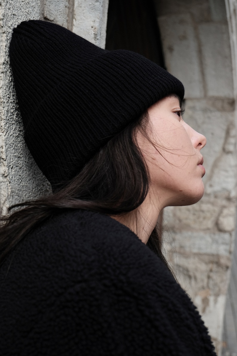 K&ouml;nnte beinhalten: Eine Frau tr&auml;gt eine schwarze M&uuml;tze und einen schwarzen Pullover. Sie schaut zur Seite und hat lange dunkle Haare.