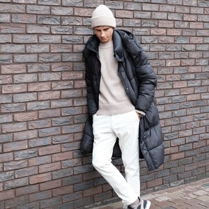 K&ouml;nnte beinhalten: Eine Person tr&auml;gt eine schwarze Daunenjacke mit Kapuze, wei&szlig;e Hose und eine beige M&uuml;tze. Die Person steht an einer Backsteinwand.