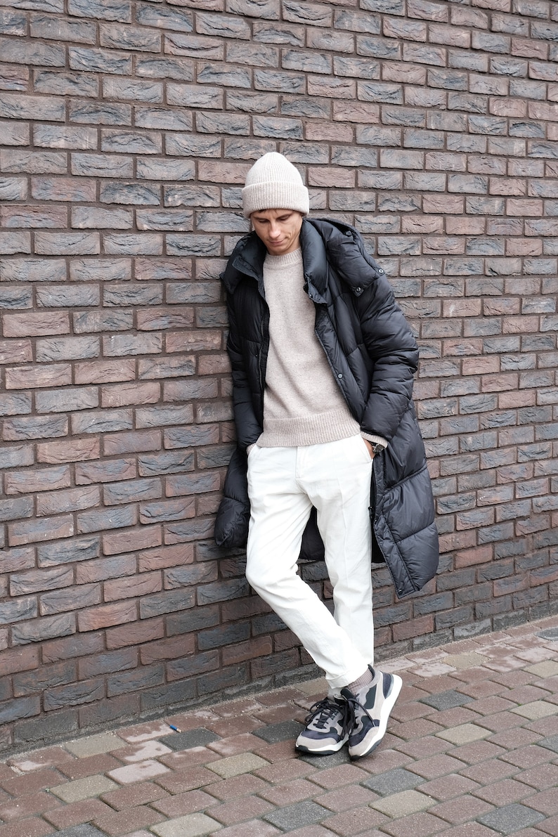 K&ouml;nnte beinhalten: Eine Person tr&auml;gt eine schwarze Daunenjacke mit Kapuze, wei&szlig;e Hose und eine beige M&uuml;tze. Die Person steht an einer Backsteinwand.