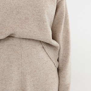 May include: A beige knitted sweater and matching pants set. The sweater has long sleeves and a relaxed fit. The pants have a wide leg and a comfortable fit.