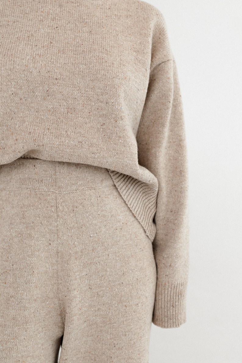 May include: A beige knitted sweater and matching pants set. The sweater has long sleeves and a relaxed fit. The pants have a wide leg and a comfortable fit.