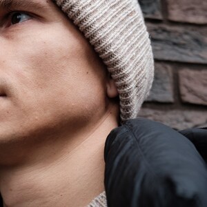 K&ouml;nnte beinhalten: Nahaufnahme einer Person, die eine beige Strickm&uuml;tze und eine schwarze Daunenjacke tr&auml;gt. Das Gesicht der Person ist teilweise sichtbar und sie schaut nach oben.