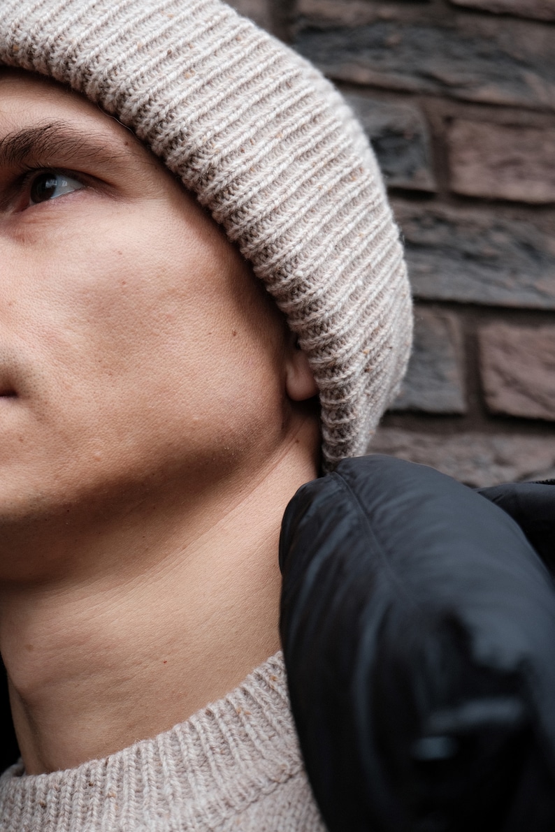 K&ouml;nnte beinhalten: Nahaufnahme einer Person, die eine beige Strickm&uuml;tze und eine schwarze Daunenjacke tr&auml;gt. Das Gesicht der Person ist teilweise sichtbar und sie schaut nach oben.