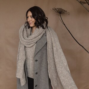 May include: A woman wearing a gray coat and a light brown scarf. The scarf is draped over her shoulders and around her neck. She is standing in front of a brown background with dried flowers.