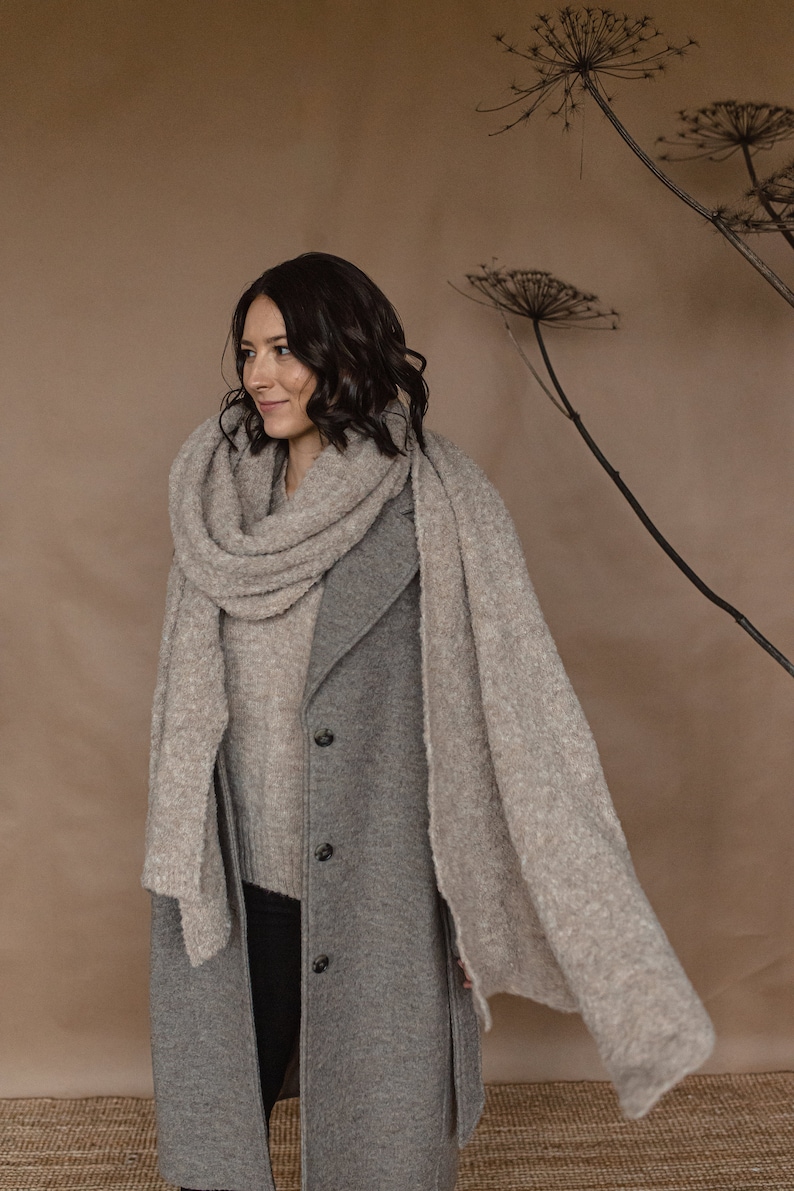 May include: A woman wearing a gray coat and a light brown scarf. The scarf is draped over her shoulders and around her neck. She is standing in front of a brown background with dried flowers.