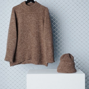 May include: A brown knitted sweater and matching beanie hat are displayed on a white platform. The sweater is hanging on a black hanger and the beanie is sitting on the platform. The background is a light blue wire mesh.