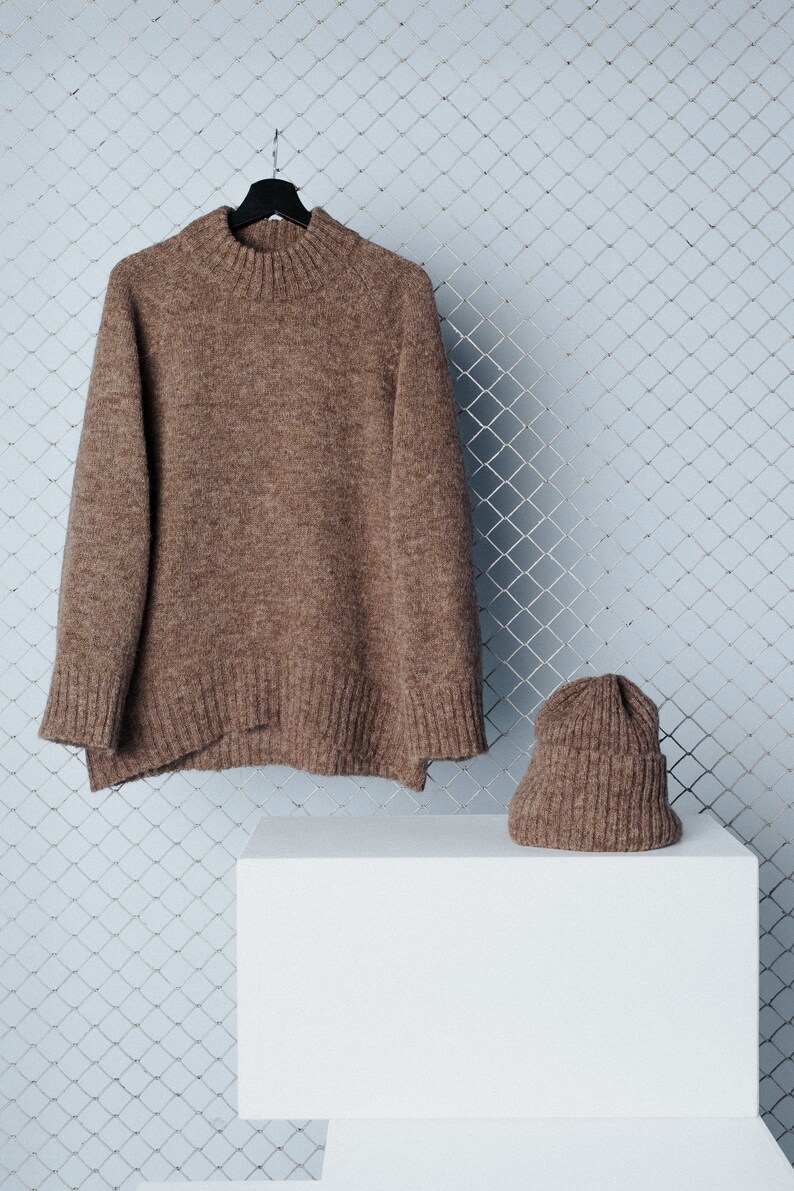 May include: A brown knitted sweater and matching beanie hat are displayed on a white platform. The sweater is hanging on a black hanger and the beanie is sitting on the platform. The background is a light blue wire mesh.