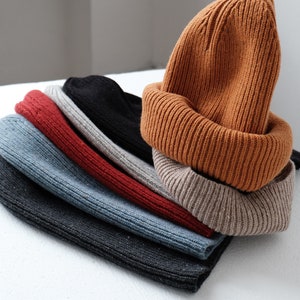 K&ouml;nnte beinhalten: Ein Stapel von sechs gestrickten M&uuml;tzen in verschiedenen Farben, darunter Schwarz, Grau, Blau, Rot, Beige und Orange. Die M&uuml;tzen sind gefaltet und &uuml;bereinander gestapelt.