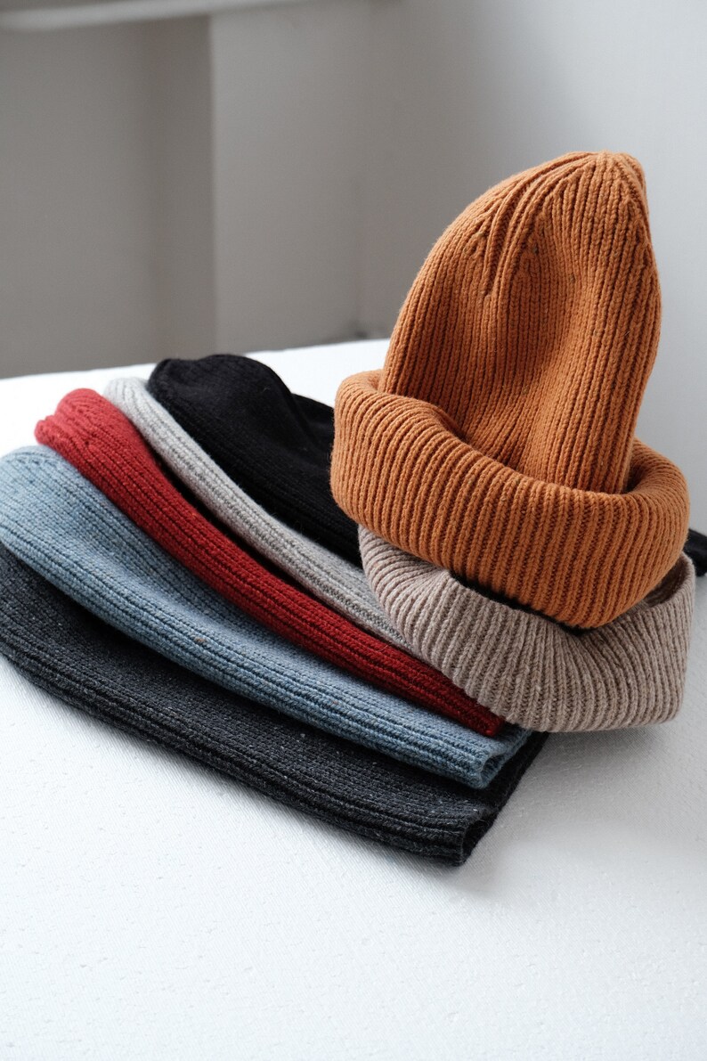 K&ouml;nnte beinhalten: Ein Stapel von sechs gestrickten M&uuml;tzen in verschiedenen Farben, darunter Schwarz, Grau, Blau, Rot, Beige und Orange. Die M&uuml;tzen sind gefaltet und &uuml;bereinander gestapelt.