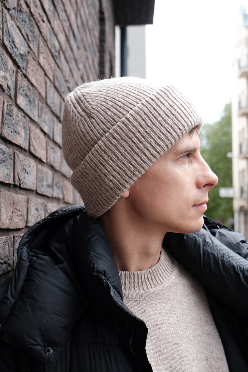K&ouml;nnte beinhalten: Ein Mann tr&auml;gt eine beige Strickm&uuml;tze und eine schwarze Daunenjacke. Er steht vor einer Backsteinmauer.