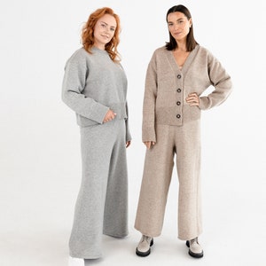 May include: Two women wearing matching knit sweater and pants sets. One set is gray and the other is beige. Both women are standing with their hands on their hips and looking at the camera.