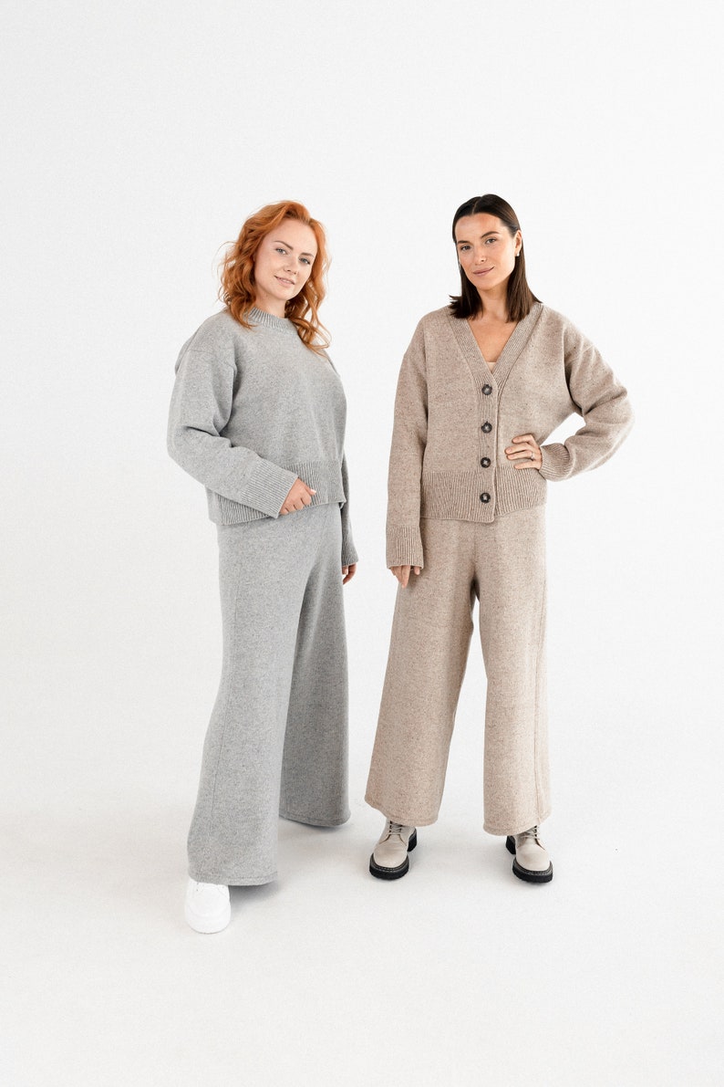 May include: Two women wearing matching knit sweater and pants sets. One set is gray and the other is beige. Both women are standing with their hands on their hips and looking at the camera.