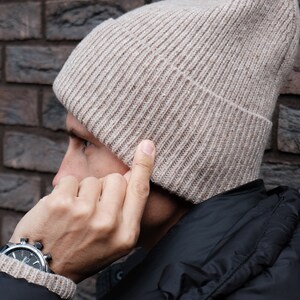 K&ouml;nnte beinhalten: Eine beige Strickm&uuml;tze mit gerippter Struktur. Die M&uuml;tze wird von einer Person getragen, die eine schwarze Jacke tr&auml;gt.