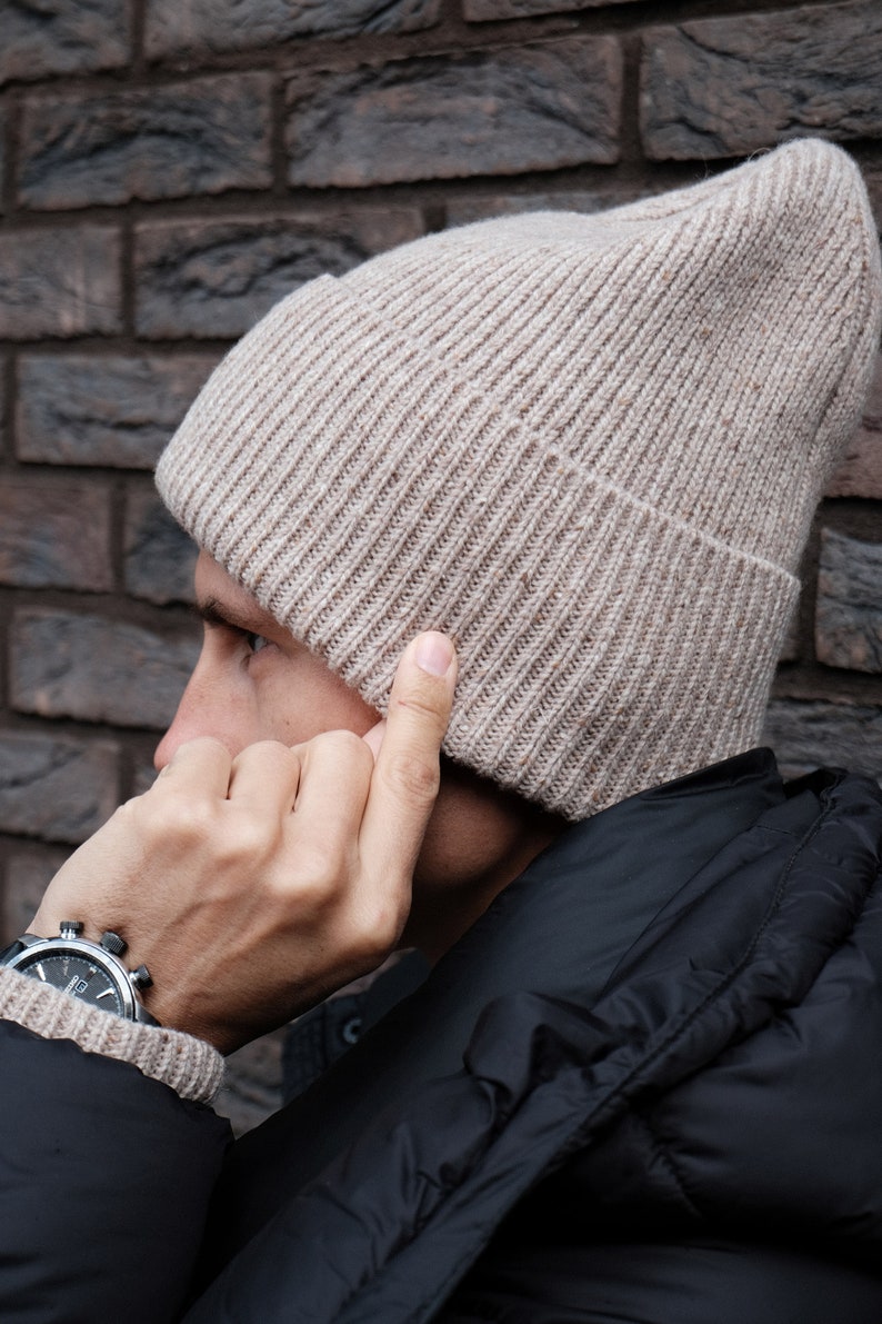 K&ouml;nnte beinhalten: Eine beige Strickm&uuml;tze mit gerippter Struktur. Die M&uuml;tze wird von einer Person getragen, die eine schwarze Jacke tr&auml;gt.