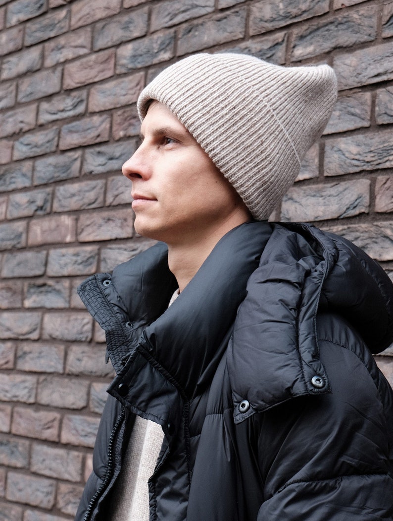 K&ouml;nnte beinhalten: Ein Mann tr&auml;gt eine beige Strickm&uuml;tze und eine schwarze Daunenjacke mit Kapuze.