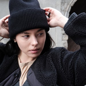 K&ouml;nnte beinhalten: Eine Frau tr&auml;gt eine schwarze M&uuml;tze und eine schwarze Fleecejacke. Die Jacke hat einen Rei&szlig;verschluss und einen Knopfverschluss. Die Frau schaut nach rechts.