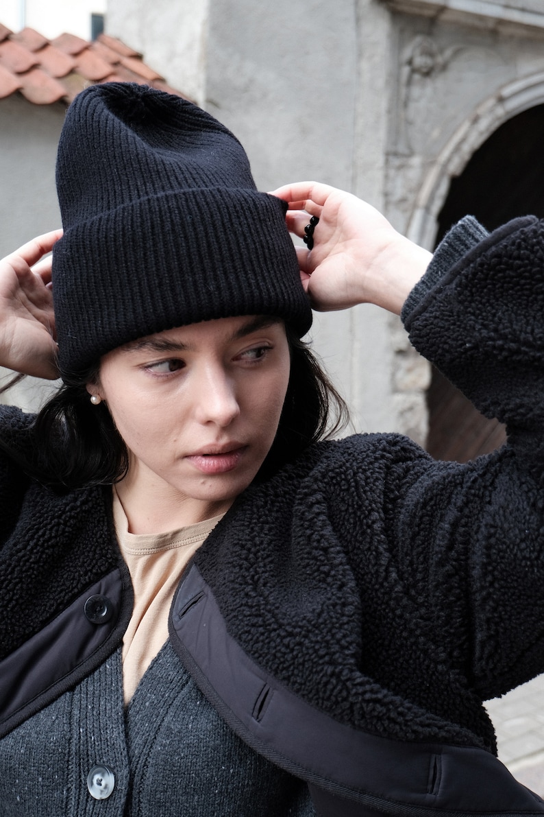 K&ouml;nnte beinhalten: Eine Frau tr&auml;gt eine schwarze M&uuml;tze und eine schwarze Fleecejacke. Die Jacke hat einen Rei&szlig;verschluss und einen Knopfverschluss. Die Frau schaut nach rechts.