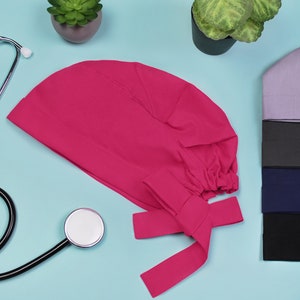 Könnte beinhalten: Eine leuchtend pinkfarbene OP-Mütze mit Schleife auf hellblauem Hintergrund. Auf der rechten Seite des Bildes liegen vier weitere OP-Mützen in verschiedenen Farben übereinander.