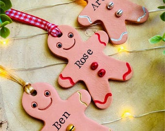 May include: Three pink gingerbread man ornaments with smiling faces, red and silver accents, and the names "Archie", "Rose", and "Ben". Each ornament has a ribbon for hanging and small bells. The ornaments are on a wooden surface.