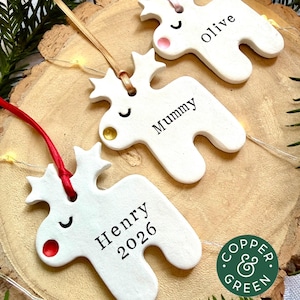 May include: Three white clay reindeer ornaments with coloured noses and ribbons. Each ornament features a name printed in black ink: Olive, Mummy, and Henry 2026. The reindeer have closed eyes and are suspended by thin satin ribbons in pink, gold, and red.