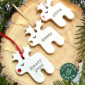 May include: Three white ceramic reindeer ornaments with star-shaped antlers, each featuring a coloured nose and a name printed in black. The ornaments are suspended by ribbons and include the text "olive," "Mummy," and "Henry 2026" on their surfaces.