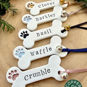 May include: Five white bone-shaped ornaments with black text reading Clover, Barkley, Basil, Waffle, and Crumble. Each ornament features a coloured paw print and a small metal bell attached by a ribbon. A green circular logo reads Copper & Green.