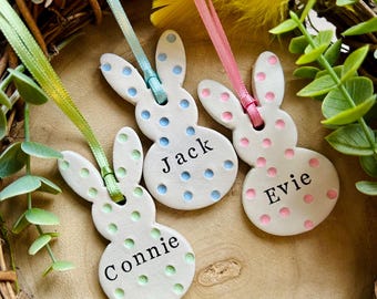 May include: Three white ceramic bunny ornaments with polka dots in blue, pink, and green. Each bunny has a name: "Connie", "Jack", and "Evie". They hang from colorful ribbons, set against a rustic wreath with floral accents.