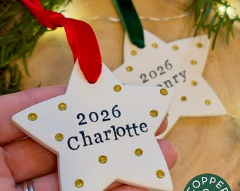 Könnte beinhalten: Zwei weiße sternförmige Keramikornamente mit goldenen Glitzerpunkten und schwarzem Text. Ein Ornament zeigt den Namen Charlotte und das Jahr 2026, das andere das Jahr 2026. Jedes Ornament hängt an einem roten oder grünen Samtband.
