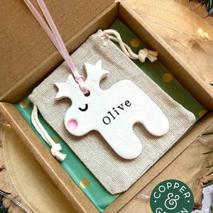 May include: A white ceramic reindeer ornament with a pink nose and the name Olive printed in black, suspended by a light pink ribbon. The ornament rests on a beige drawstring fabric pouch inside a brown cardboard gift box with green paper.