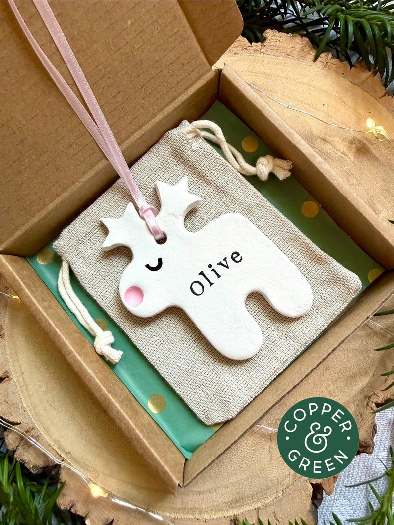 May include: A white ceramic reindeer ornament with a pink nose and the name Olive printed in black, suspended by a light pink ribbon. The ornament rests on a beige drawstring fabric pouch inside a brown cardboard gift box with green paper.