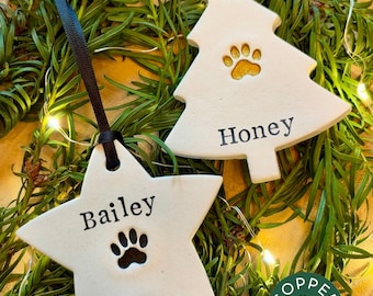 May include: Two white ceramic ornaments, one star-shaped and one tree-shaped, hanging by ribbons. The star ornament features the name Bailey and a black paw print. The tree ornament features the name Honey and a gold paw print.