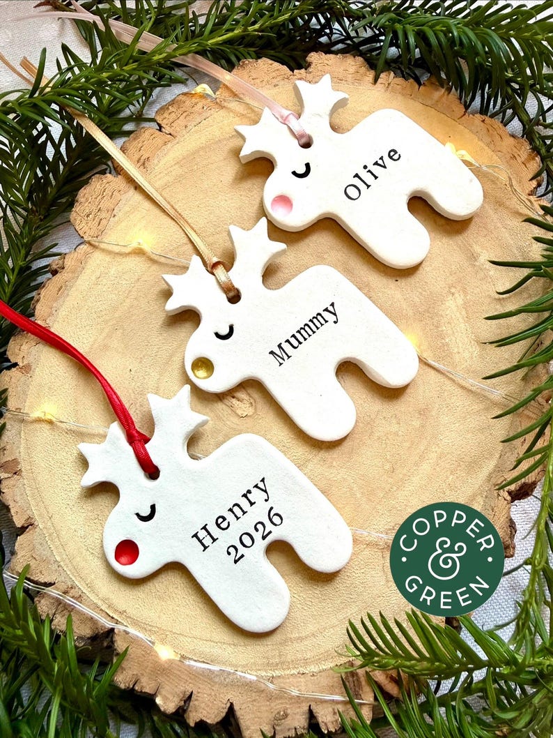 May include: Three white ceramic reindeer ornaments with star-shaped antlers, each featuring a coloured nose and a name printed in black. The ornaments are suspended by ribbons and include the text "olive," "Mummy," and "Henry 2026" on their surfaces.