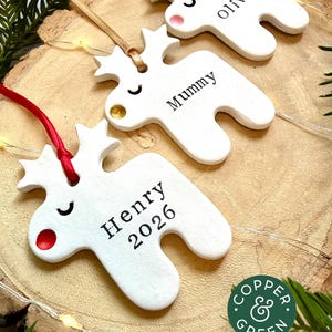 May include: Three white reindeer-shaped ornaments made of clay, each featuring a closed-eye expression and a colored nose. The ornaments are personalized with the names Olive, Mummy, and Henry 2026, and hang from ribbons in pink, gold, and red.