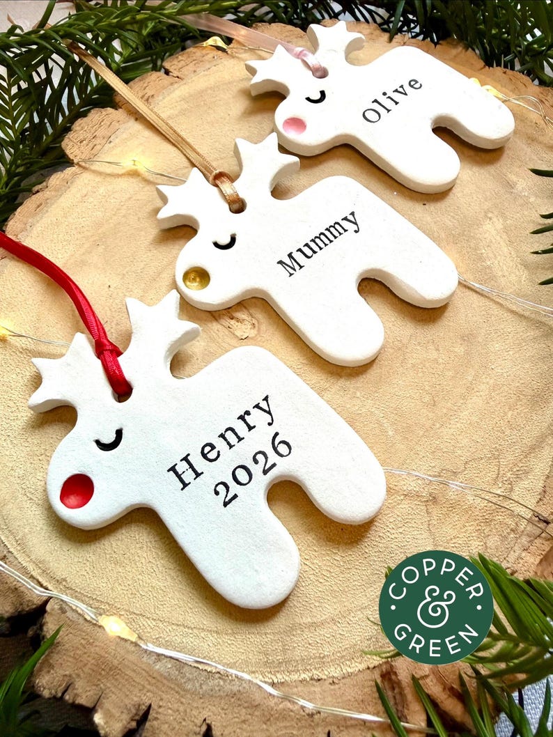 May include: Three white reindeer-shaped ornaments made of clay, each featuring a closed-eye expression and a colored nose. The ornaments are personalized with the names Olive, Mummy, and Henry 2026, and hang from ribbons in pink, gold, and red.