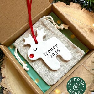 May include: A white ceramic reindeer ornament with a red nose and closed eyes, featuring the text Henry 2026. The ornament hangs from a red ribbon and is placed on a beige drawstring fabric pouch inside a cardboard gift box with green paper.