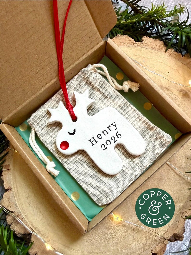 May include: A white ceramic reindeer ornament with a red nose and closed eyes, featuring the text Henry 2026. The ornament hangs from a red ribbon and is placed on a beige drawstring fabric pouch inside a cardboard gift box with green paper.