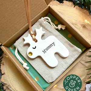 May include: A white ceramic reindeer ornament with a gold nose and the word Mummy printed in black, hanging by a peach ribbon. The ornament rests on a beige drawstring fabric pouch inside a cardboard gift box with green polka dot paper.