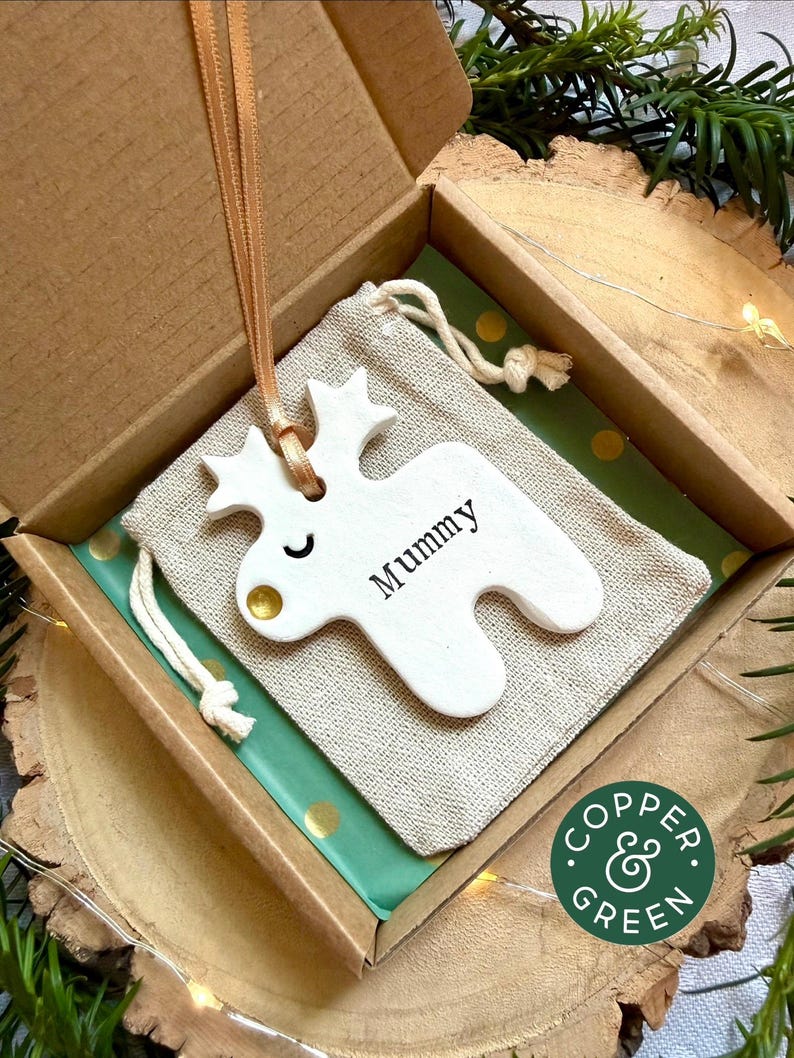 May include: A white ceramic reindeer ornament with a gold nose and the word Mummy printed in black, hanging by a peach ribbon. The ornament rests on a beige drawstring fabric pouch inside a cardboard gift box with green polka dot paper.