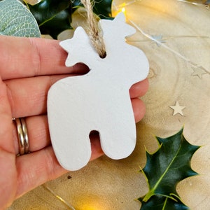 May include: A white, matte-finished clay ornament shaped like a stylized reindeer with a star on its head. The ornament features a hole at the top threaded with a natural jute twine loop for hanging, providing a simple, neutral aesthetic.