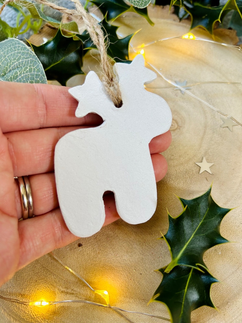 May include: A white, matte-finished clay ornament shaped like a stylized reindeer with a star on its head. The ornament features a hole at the top threaded with a natural jute twine loop for hanging, providing a simple, neutral aesthetic.