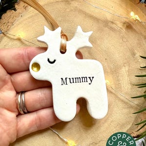 May include: A white ceramic reindeer ornament with a gold-painted nose and a closed eye. The word "Mummy" is printed in black on the body. A tan ribbon is threaded through a hole in the top, between the antlers, for hanging the decoration.