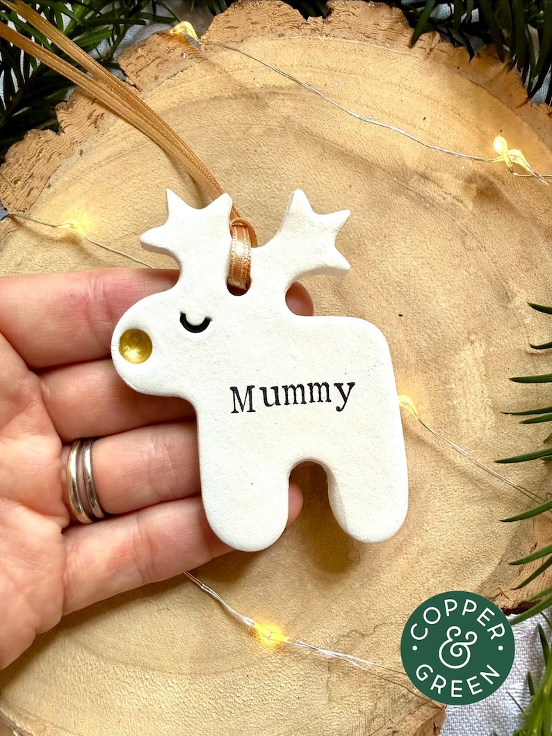 May include: A white ceramic reindeer ornament with a gold-painted nose and a closed eye. The word "Mummy" is printed in black on the body. A tan ribbon is threaded through a hole in the top, between the antlers, for hanging the decoration.
