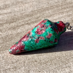 May include: A gemstone pendulum with a silver chain. The pendulum is a cone shape, with a vibrant mix of green and red colors. The stone has a textured surface, and the chain is attached at the top.