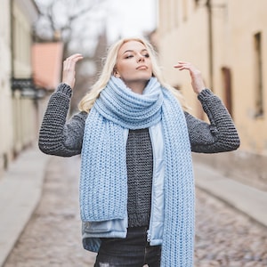 May include: A woman wearing a gray sweater, a blue knitted scarf, and blue jeans. She is standing in a cobblestone street.