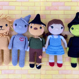 May include: Five crocheted dolls representing characters from The Wizard of Oz. The dolls include the Lion, Tin Man, Scarecrow, Dorothy, and the Wicked Witch. Each doll is handmade with detailed features and vibrant colors, standing against a yellow brick wall.