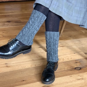 May include: Gray knitted leg warmers with a cable knit design are worn over black tights, paired with black leather shoes. The leg warmers add a touch of warmth and style to the outfit.